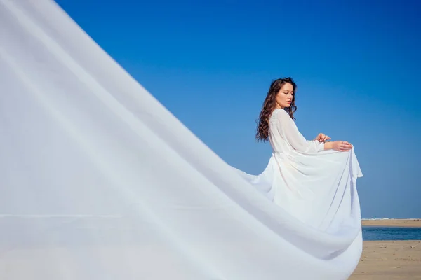 Schöne brünette Frau in weißem Kleid mit langer Schleppe an einem Strand, der Einsamkeit und Freiheit genießt — Stockfoto