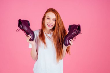 Kızıl saçlı, pamuklu tişörtlü kadın pembe stüdyoda bordo bağcıklı botlar giyiyor.