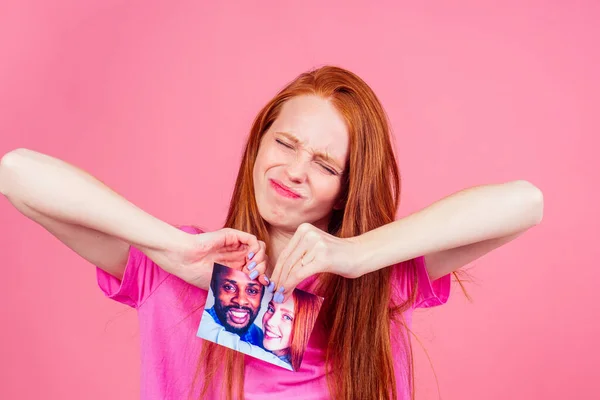 Rothaarige Ingwerfrau im rosafarbenen Studiohintergrund zerreißt Fotokarte lächelnden internationalen Paares das Ende der Beziehung — Stockfoto
