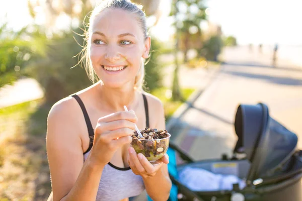 Mulher loira segurando tapete de ioga e sorvete de iogurte vegan com chocolate de feijão de cacau e amendoim em fundo tropical — Fotografia de Stock