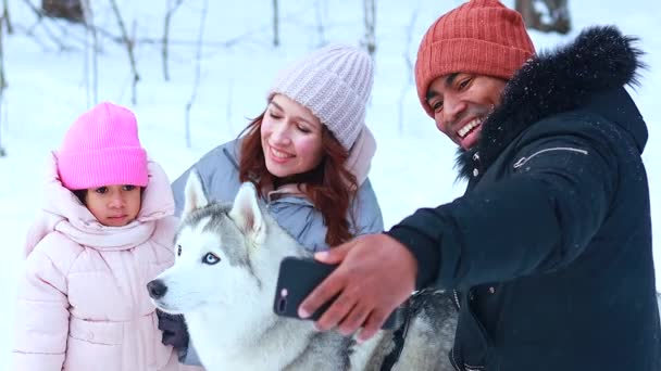 Afro człowiek ze swoją białą żoną zabawia się z piękną córką grając husky w zaśnieżonym parku robiąc selfie na telefon aparatu — Wideo stockowe