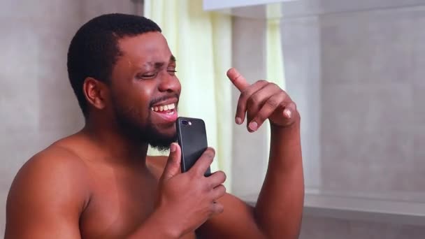 Atrakcyjny młody wesoły mężczyzna śpiewa podczas mycia pod prysznicem, trzymając telefon — Wideo stockowe