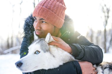 Karışık ırk adamı kış parkında köpeğine sarılıyor.