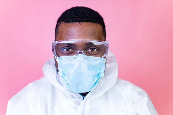 Cientista afro-americano em uniforme de suíte PPE mostrando remédio líquido frasco de vacina frasco coronavírus estúdio parede rosa — Fotografia de Stock