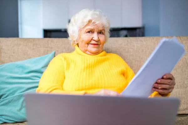 happy old person looking at paper document and laptop sitting on sofa in living room