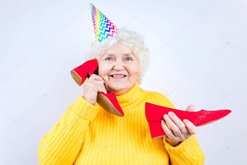 older woman with a gift wear yellow sweater and horn cap on a white background holding red high heels shoes