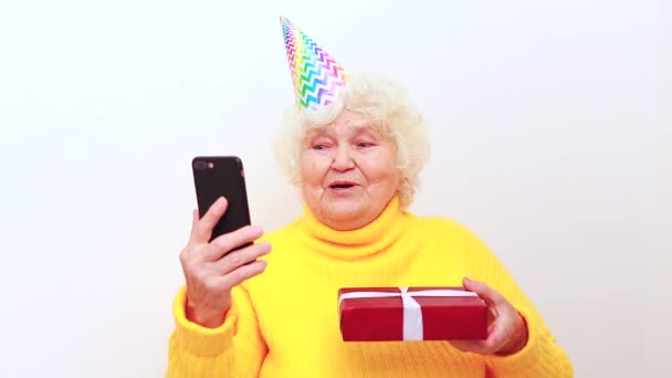 Donna anziana con un regalo indossare maglione giallo e cappuccio corno su uno sfondo bianco che tiene smartphone — Video Stock