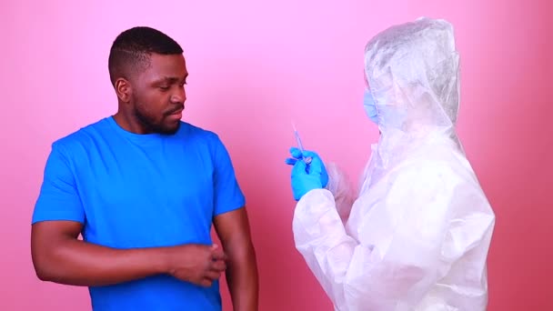 Африканский американец ученый в униформе СИЗ, показывающий лекарство флакон с жидкой вакциной коронавируса студия розовая стена — стоковое видео