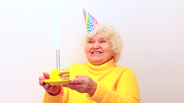 Older woman with a gift wear yellow sweater and horn cap on a white background holding plate with cake with fireworks — Stock Video
