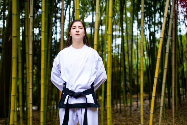 Uma jovem mulher com um cinto preto treinadores em arte marcial ao ar livre fundo de bambu — Fotografia de Stock