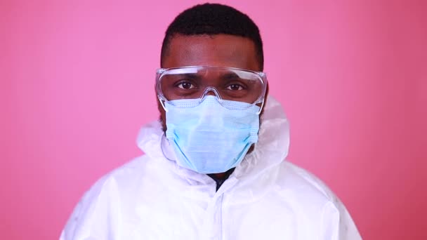 Uomo scienziato afroamericano in uniforme PPE suite che mostra medicina vaccino liquido flacone coronavirus studio parete rosa — Video Stock