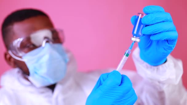 Cientista afro-americano em uniforme de suíte PPE mostrando remédio líquido frasco de vacina frasco coronavírus estúdio parede rosa — Vídeo de Stock