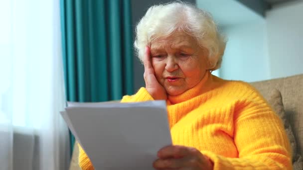 Shoked amazed old woman white gray-haired sitting on the sofa in living room with bills — Stock Video