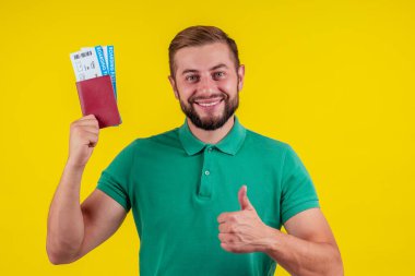 Güneş gözlüklü ve yeşil tişörtlü mutlu bir gencin portresi sarı arka planda izole edilmiş bir pasaport taşıyor.