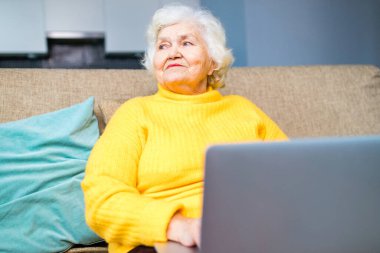 Yaşlı gri saçlı kadın oturma odasında otururken gözlerini kaçırıyor.