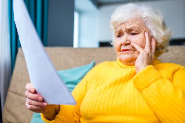 Şok olmuş yaşlı kadın beyaz saçlı oturma odasındaki kanepede dizüstü bilgisayar ve faturalarla oturuyor.