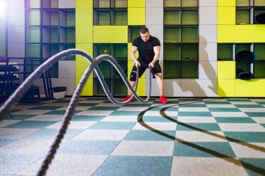 Genç ve yakışıklı erkek sporcu vücut kasları için egzersiz yapıyor. Spor salonunda sarı arka planda savaş halatları var.