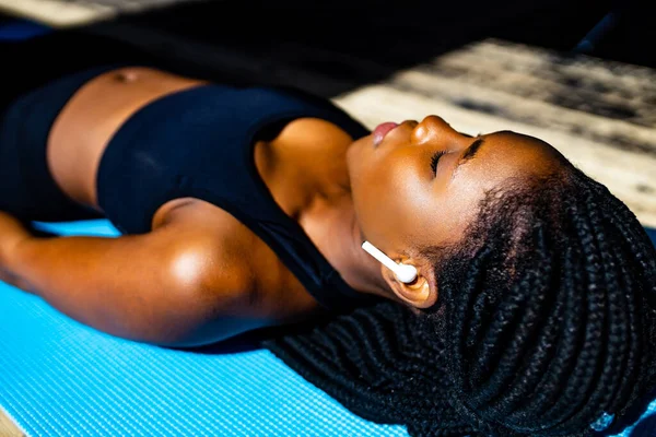 Mulher africana americana em sutiã desportivo preto está deitado no chão tapete de ioga olhos fechados e ouvir um mantra de áudio meditar — Fotografia de Stock