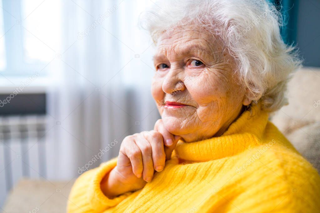 Thoughtful senior lady sitting at home with her fingers to her chin in living room