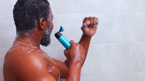 Aantrekkelijke jonge vrolijke man zingen tijdens het wassen in de douche, met shampoo fles — Stockvideo