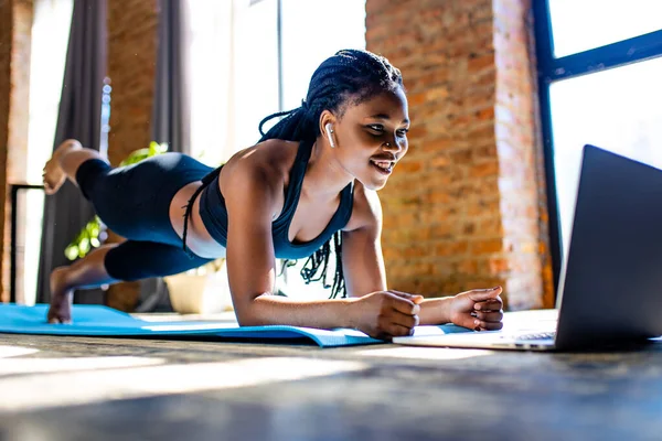 Blandad ras frisk kvinna i svart sportig kostym under hennes fitness träning hemma mot bakgrund av panoramafönster solljus — Stockfoto