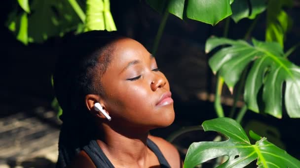 Africano americano donna in nero sportivo reggiseno è seduto loto posa su yoga mat pavimento chiuso gli occhi e ascoltare un audio meditare mantra mani namaste — Video Stock