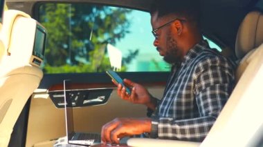 Yakışıklı, başarılı, Afro-Amerikan iş adamları şık takım elbiseli, lüks bir arabada oturuyor ve dizüstü bilgisayarla çalışıyor.