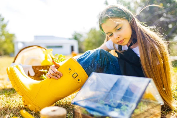 Gadis sekolah yang cantik menghabiskan waktu di luar ruangan di taman mengenakan sepatu karet kuning melihat ke dalam sebuah rumah kaca kecil — Stok Foto