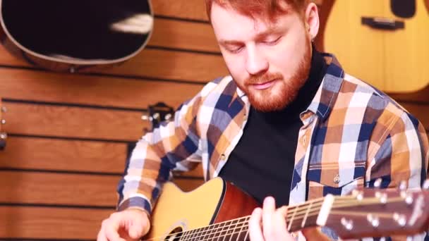 Ier roodharige bebaarde knappe man in bruin geruit casual shirt kiezen van een gitaar in een muziekwinkel — Stockvideo