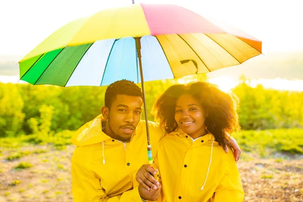Africano americano esposa y marido en amarillo impermeable con un capucha sensación feliz al atardecer al aire libre verano bajo arco iris paraguas — Foto de Stock