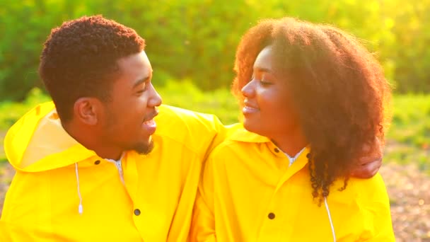 Αφροαμερικάνα σύζυγος και σύζυγος με κίτρινο αδιάβροχο με κουκούλα που αισθάνονται ευτυχισμένοι στο ηλιοβασίλεμα στην ύπαιθρο το καλοκαίρι μιλώντας — Αρχείο Βίντεο