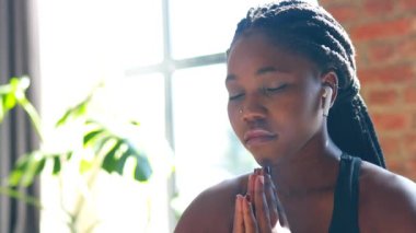 Siyah spor sutyen giyen Afrikalı Amerikalı kadın yoga minderinde poz veriyor gözleri kapalı ve sesli meditasyon mantra elleri namaste dinliyor.