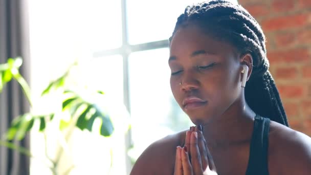 Afrikansk amerikansk kvinna i svart sportig behå sitter lotus poserar på yogamatta golv stängda ögon och lyssnar på en ljud meditera mantra händer namaste — Stockvideo