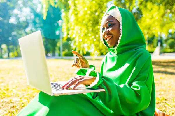 Латиноамериканка в зеленом мусульманском хиджабе с ярким макияжем и пирсингом носа, работающая на ноутбуке, сидит на траве в летнем парке — стоковое фото
