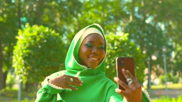 Wielokulturowa kobieta w zielonej sukience muzułmańskiej z jasnym makijażem i przeszywającym nosem robiąca selfie na świeżym powietrzu latem . — Wideo stockowe