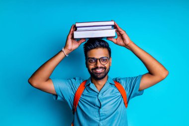 Hintli öğrenci kameraya baktığı için mutlu. Mavi stüdyoda gözlük ve turuncu sırt çantası takıyor.