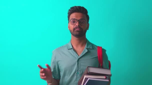 Étudiant indien homme se sentant heureux en regardant caméra porter des lunettes et sac à dos orange et en disant sur les livres cognitifs en studio bleu — Video
