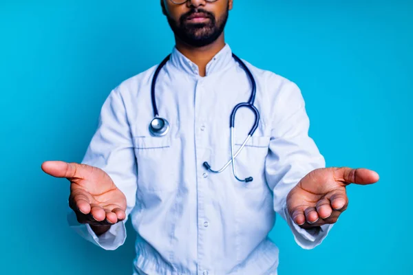 Pakistański lekarz w białym płaszczu ze stetoskopem wskazującym na przestrzeń do kopiowania z ręczną makietą na tle niebieskiego studia — Zdjęcie stockowe