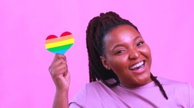 Çekici bir İspanyol kadının portresi pembe stüdyoda LGBT gökkuşağı kalbi tutuyor.