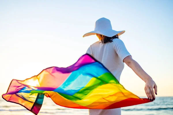 Mujer ondeando con la bandera del arco iris y sombrero en la playa al atardecer — Foto de Stock