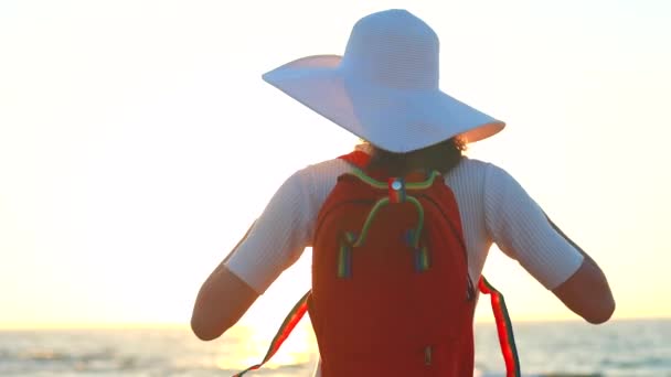 Mulher de vestido branco e chapéu de palha com mochila laranja olhando para o lado de trás do mar — Vídeo de Stock