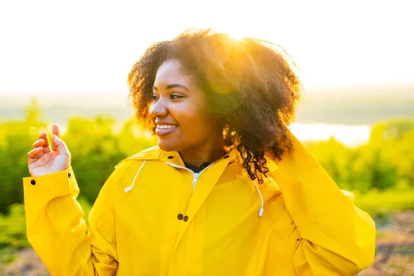 Afro-américaine en imperméable jaune avec une capuche se sentant heureux au coucher du soleil en plein air été — Photo