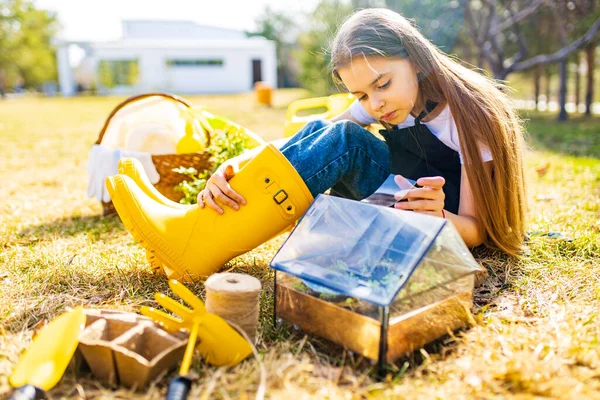 Gadis sekolah yang cantik menghabiskan waktu di luar ruangan di taman mengenakan sepatu karet kuning melihat ke dalam sebuah rumah kaca kecil — Stok Foto