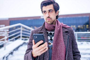 Havalı kahverengi ceketli sakallı adam dışarıda akıllı telefon tutuyor.