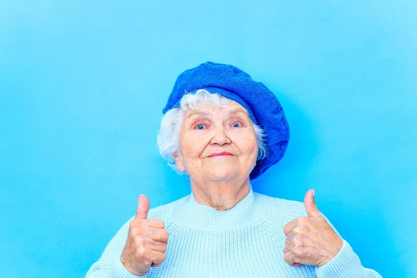 Vacker pensionerad kvinna i blå basker och varm pullover tittar på kameran och ler i blå vägg bakgrund i studio — Stockfoto