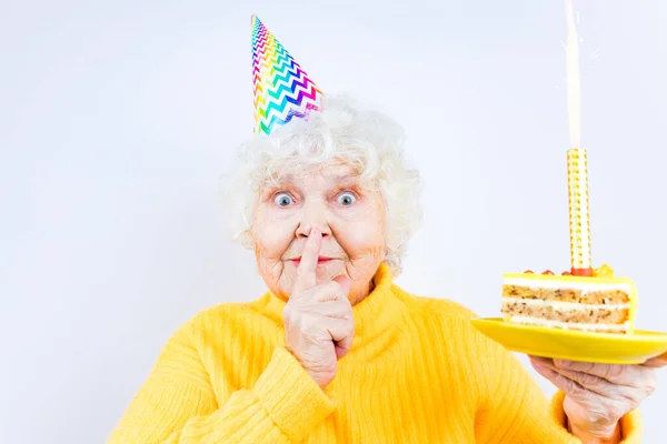 Older woman with a gift wear yellow sweater and horn cap on a white background holding plate with cake with fireworks — Stock Photo, Image