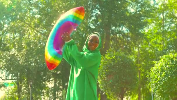 Mujer afro moderna en hijab verde con maquillaje brillante y nariz perforante hokding globo de aire del arco iris en pak de verano al aire libre. — Vídeo de stock