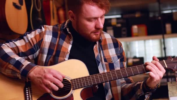 Irlandês ruivo barbudo bonito homem em marrom xadrez casual camisa escolher uma guitarra em uma loja de música — Vídeo de Stock