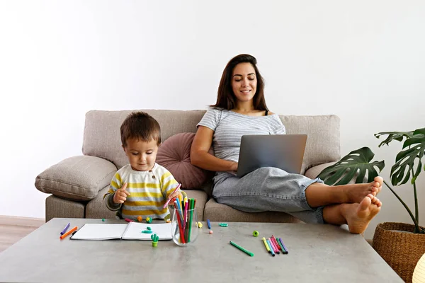 Young beautiful woman multitasking, working from home & teaching two year old son to draw. Woman spending quality time with her toddler child. Homeschooling concept. Close up, copy space, background.