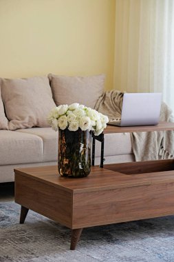 Modern bej tekstil kanepesi ve ahşap desenli masa üzerindeki dizüstü bilgisayarlı oturma odası, doğal ışık. Evin iç kısımlarında ev bitkileri. Arkaplan, boşluğu kopyala /